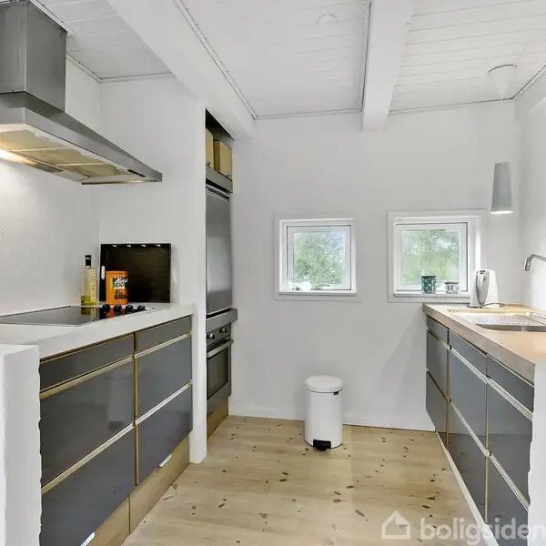 Køkken med mørkegrå skabe, indeholdende en vask og komfur, i aktion med madlavning. Rummet er lyst med træloft og gulv, og to små vinduer lader naturligt lys ind.