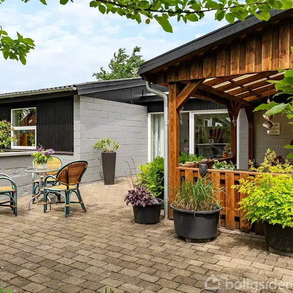 Terrasse med flisebelægning, hvor havemøbler står ved et bord. Træpergola fyldt med grønne planter. Omgivet af et hus malet i sort og grå med en frodig have.