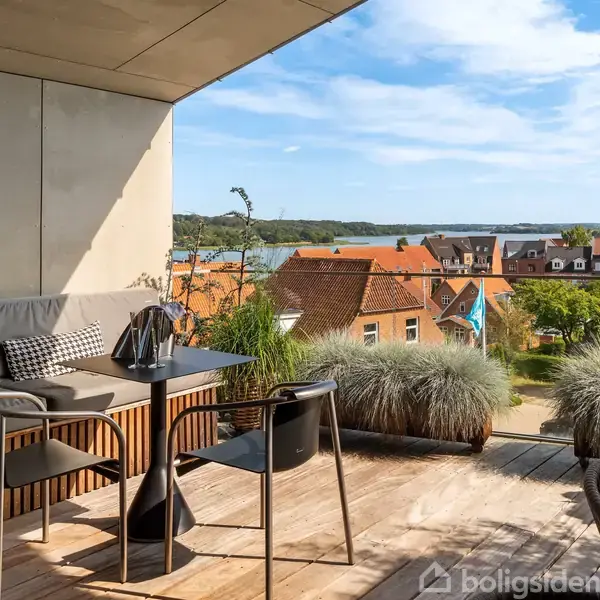 En terrasse med et moderne bord og stole, der står på et trægulv. Baggrunden består af huse med røde tage og en sø med grønne områder.