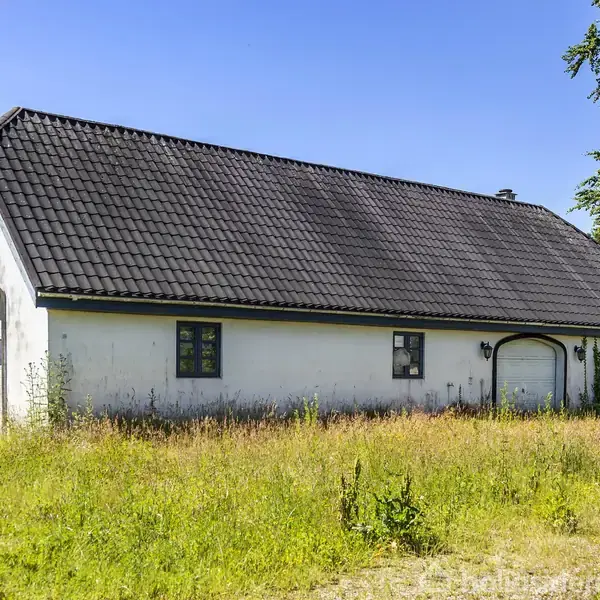 En hvid bygning med sort tag står på en græsbevokset grund. Der er to portåbninger og flere vinduer. Sollyset skinner klart, og et træ er synligt i baggrunden.