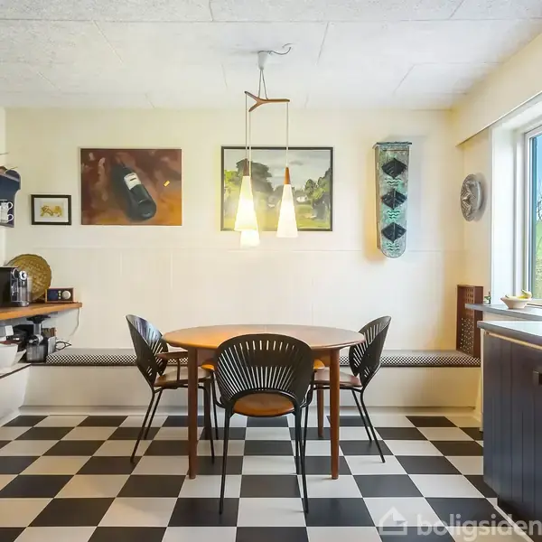 Rundt spisebord omgivet af sorte stole står på et skakternet gulv. En sort brændeovn til venstre. Lyse lamper hænger over bordet. Væggen har kunst og vindue til højre.