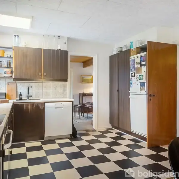 Køkken med sorte og hvide skakternet gulv, mørke træskabe, opvaskemaskine og køleskab. Bordplader er hvide. Der er åbent til en spisestue i baggrunden.