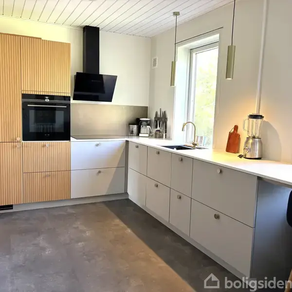 Et moderne køkken med trælåger, en indbygget ovn og emhætte. Køkkenbordet er hvidt med en blender og skærebræt. Store vinduer giver naturligt lys.