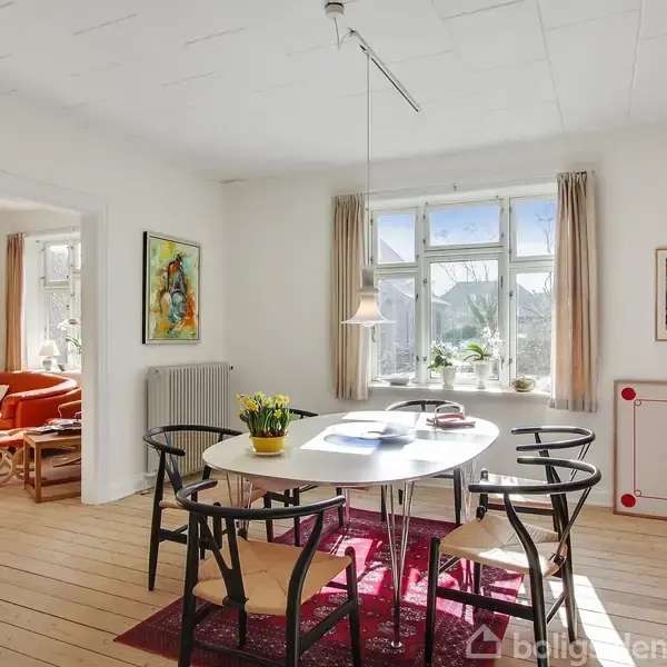 Et ovalt spisebord står i et lyst rum med seks sorte stole omkring det. På bordet ligger bøger, og en vase med blomster. Baggrunden viser et vindue og andre møbler.