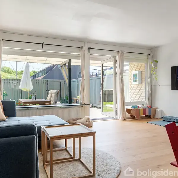 Stue med blå sofa og sofabord, vendt mod udendørs terrasse gennem stor glasdør. Legetøj i hjørnet, fjernsyn på væggen, lyse trægulve og store vinduer med gardiner.