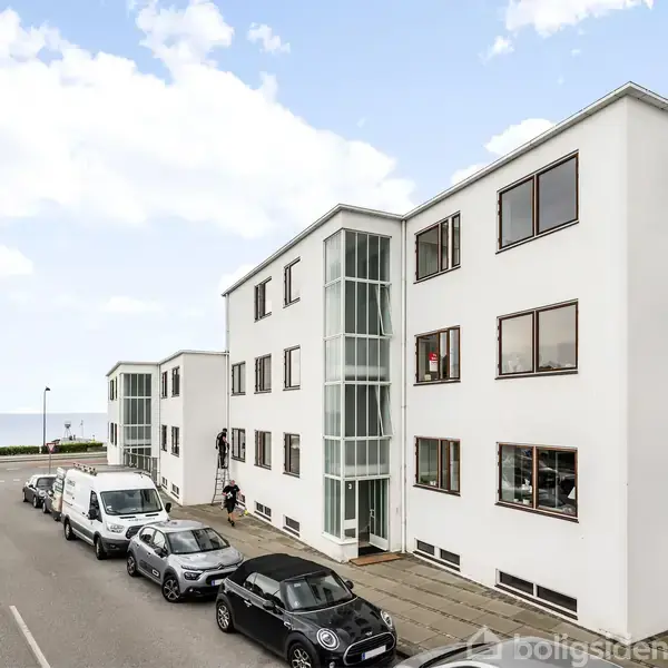 Hvidt lejlighedskompleks langs vejen, mens biler holder parkeret foran. I baggrunden ses havudsigt og blå himmel.