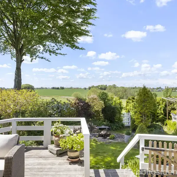 Terrasse med havemøbler og planter står i sollys. I baggrunden ses en have med træer og buske mod en åben mark og blå himmel med skyer.