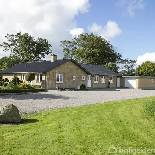 Huset står stille i et grønt landskab med græsplæne og træer. Det er et enetages byggeri med garage, placeret på en grusbelagt vej.