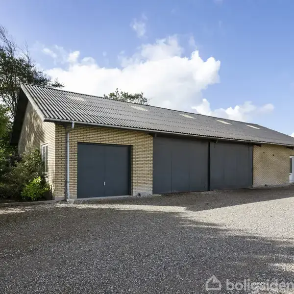 En stor, beige murstensbygning med tre garagedøre står stille i en grusbelagt indkørsel, omgivet af træer under en blå himmel.