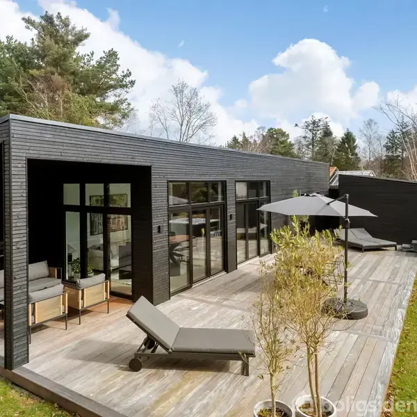 Sort, moderne hus med store vinduer åbner ud mod en træterrasse. Terrassemøbler og parasol giver udendørs hygge. Omgivet af græsplæne og høje træer under en blå himmel med skyer.