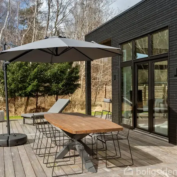 En træterrasse med et stort spisebord under en parasol. Bordet omgivet af moderne stole. Baggrunden består af en grøn have og en sort væg med store vinduer.