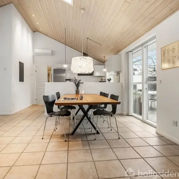 En moderne stue med en grå sofa står i hjørnet. Ved siden af er der to runde sofaborde. Et stort fladskærms-tv hænger på væggen. Store vinduer giver udsigt til haven.