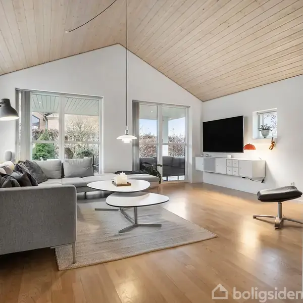 Gråt sofaarrangement står i stue med trægulv og højt loft. Tv på væggen, runde borde på tæppe. Store vinduer giver udsigt til have. Lænestol ved siden af.