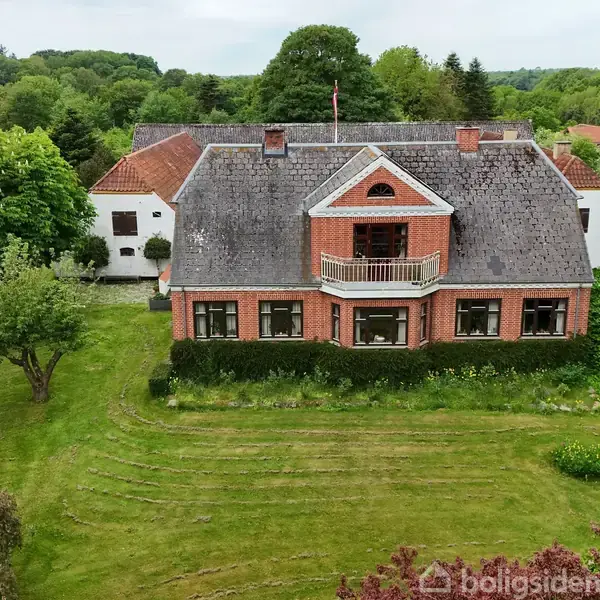 Et stort murstenshus med en balkon i midten, omkranset af en velplejet have med buske og træer. Det ligger i et skovområde.