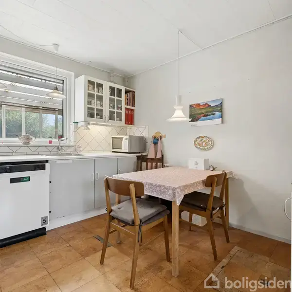 Et køkkenbord med en dug og to stole står ved en væg. Et vindue, opvaskemaskine og overskabe er placeret omkring. Indretningen er lys og enkel. En lampe hænger over bordet.