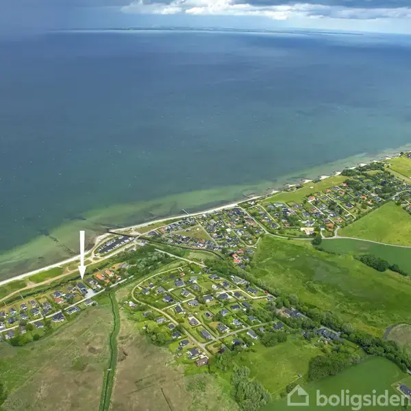 Kystnært byområde set fra luften, med markerede huse og veje langs kystlinjen. Havet strækker sig ud til horisonten, mens grønne marker omkranser beboelsesområderne.