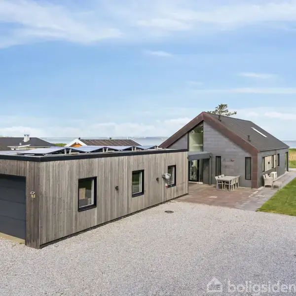 Moderne hus med træbeklædning, står stille på en grusplads med havudsigt i baggrunden. Huset har store vinduer og en terrasse med stole, omgivet af græsplæne og naboer.