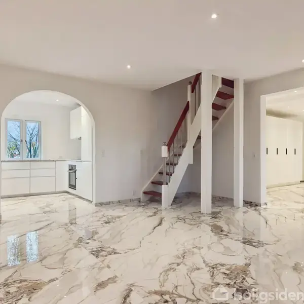 Stue med glat marmorgulv, trapper til første sal, og en bueåbning til hvidt køkken i baggrunden, under nedhængte spotlights.