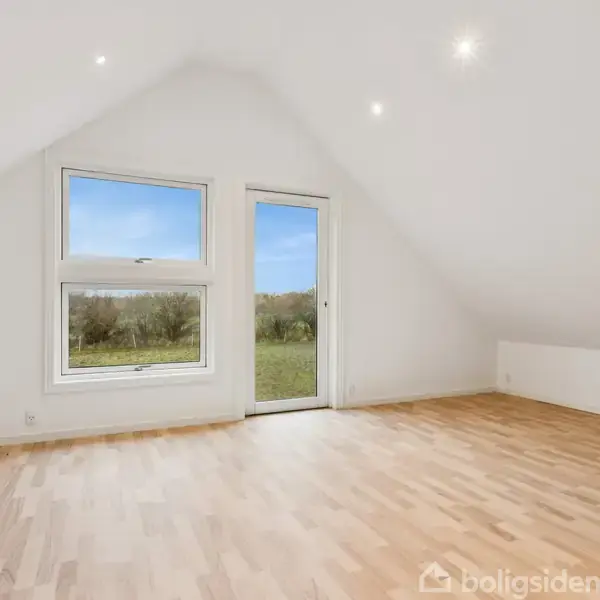 Et rummeligt, lyst rum med trægulv; vindue og dør leder udenfor mod en grøn have. Rummet er tomt og har en hvid væg og skrå loftlinjer.