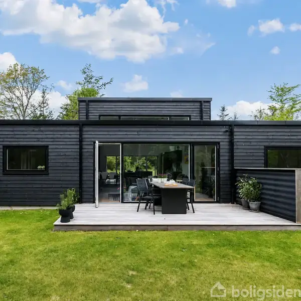 Et moderne, sort hus omgivet af tæt skov står midt i et grønt landskab. I baggrunden ses en flad horisont med skyer og blå himmel.