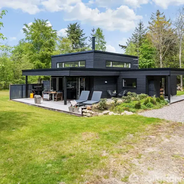 Sort, moderne sommerhus omkranset af græsplæne, træer og grusbelagt indkørsel. En terrasse med havemøbler og liggestole ses foran huset. Træer danner en naturskøn kulisse bag huset.