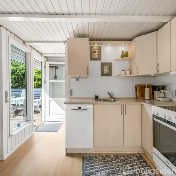 Køkken med hvide skabe og indbygget ovn. En vask ses ved siden af kaffemaskine og brødrister. Terrasse med havemøbler udenfor glasskydedør. Lyst træ på gulv og loft.