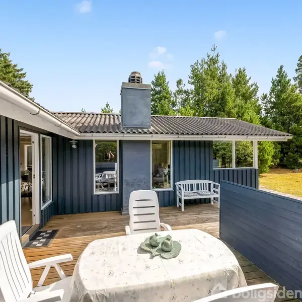 Terrassebord med hvide stole står på en træterrasse ved et blåmalet sommerhus, omgivet af græsplæne og træer. En dør fører indenfor, og himlen er klar og blå.