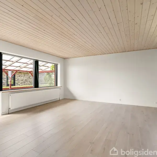 Stue med lyse trægulve og træloft. Store vinduer til venstre giver udsigt til en overdækket terrasse med røde træbjælker. Radiatorer under vinduerne. Ingen tekst fundet.
