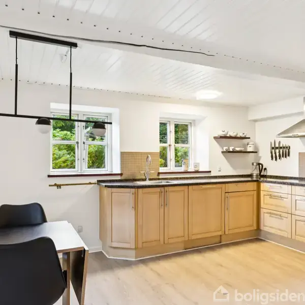 Køkken med træskabe og sort bordplade, har integreret emhætte og hvide vægge. To vinduer lader naturligt lys ind. Et bord med sorte stole står ved siden af.