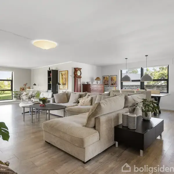 Stue med beige hjørnesofa og sofabord står centralt. Store vinduer i baggrunden giver lysindfald, med planter og billeder på væggene. Spisebord og lamper ses i baggrunden.