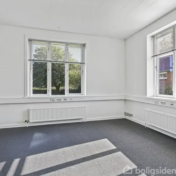 Et tomt værelse har tæppebelagt gulv og hvide vægge med store vinduer, der giver udsigt til en grøn have og en murstensbygning. Sollys kaster skygger på gulvet.