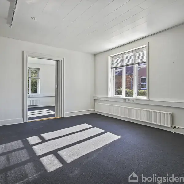 Et tomt rum med hvidmalede vægge er oplyst af sollys gennem et vindue. Skygger falder på det mørke gulvtæppe. En døråbning fører til et andet rum.