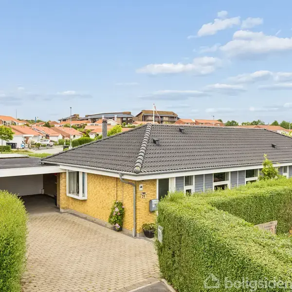 Et gult murstenshus med mørkegråt tag står stille i et forstadsområde, omgivet af en velplejet hæk. En brolagt indkørsel fører til en carport, mens naboernes tage er synlige i baggrunden.