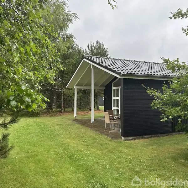 Træhus med terrasse, omgivet af grønne træer og græsplæne. Et sæt havemøbler står under en overdækket del af terrassen. Himlen er overskyet.