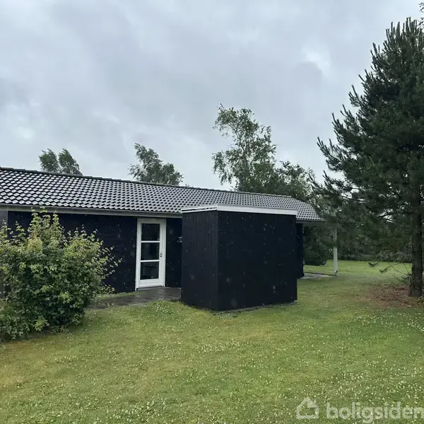 Et sort træhus står stille omgivet af grønne buske og græsplæne, med høje træer i baggrunden under en overskyet himmel.