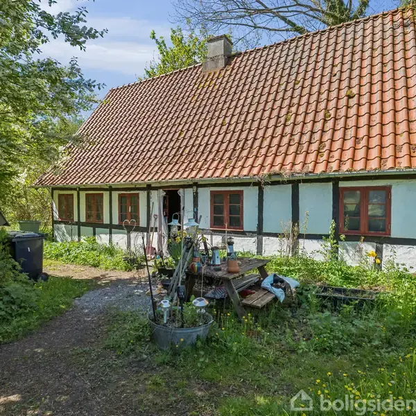 Et gammelt bindingsværkshus står stille omgivet af frodig vegetation og store træer. Tiden ser ud til at have efterladt et par redskaber og en skraldespand nær indgangen.