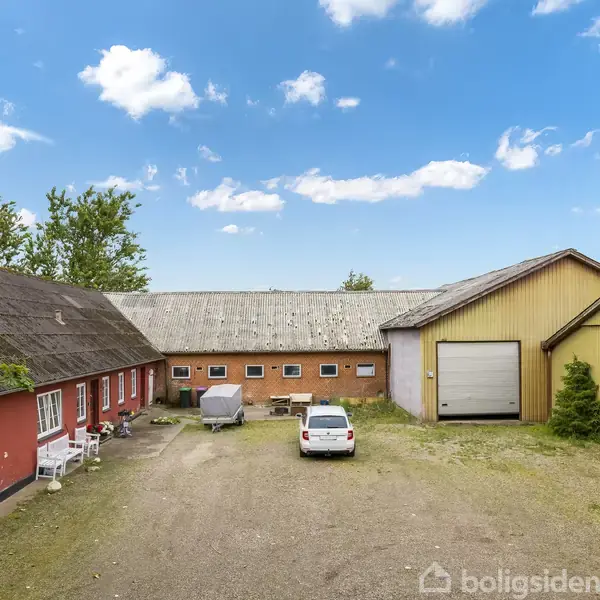 Rød og gul ladebygning står stille på en gårdsplads med grus, omgivet af træer og blå himmel. Midt på pladsen holder en hvid bil ved siden af en trailer.