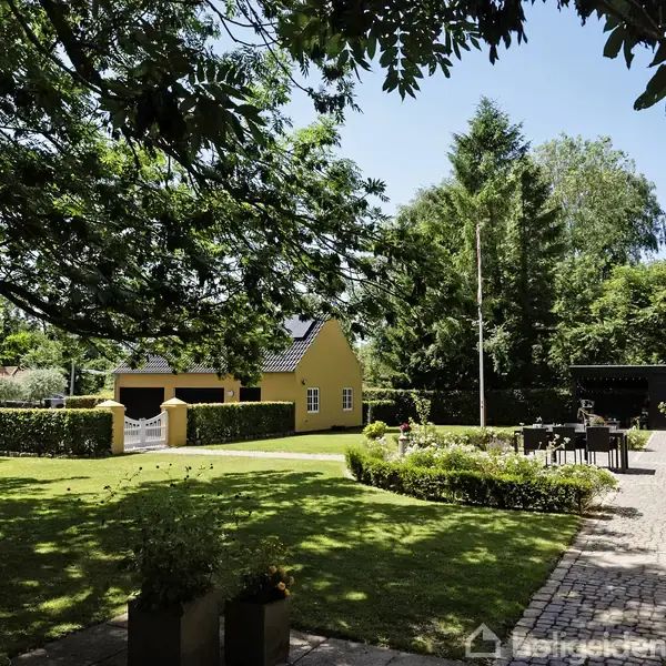 En gul bygning står i en frodig have med buske og træer. Der er en lade og et terrasseområde med havemøbler i sollys.
