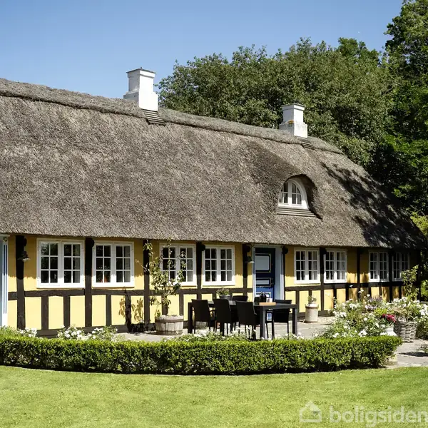 Stråtækt bindingsværkshus med blå døre og vinduer står i en frodig have med grønne plæner og blomsterkrukker, omgivet af træer under en klar blå himmel.