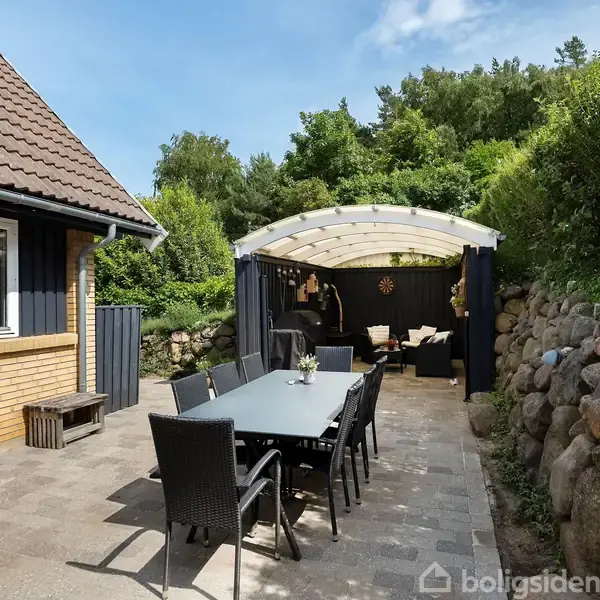 Terrasse med langt spisebord og stole under halvtag; omgivet af klippevæg og have. Der ses en åben stue i baggrunden med sofa og planter.