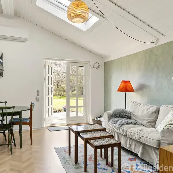 Stue med sofa og bord. En lampe lyser rummet, som åbner op via franske døre til en have. Skandinavisk indretning med træstol, grøn væg og lyse farver.