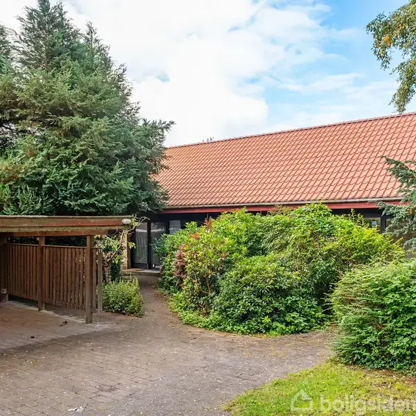 En villa med rødt tegltag, omgivet af tæt grøn beplantning og træer, står ved siden af en carport i træ. Flisebelagt indkørsel fører til huset.