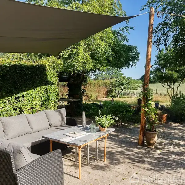 En grå udendørs sofa står på en flisebelagt terrasse med et solsejl over. Omgivet af grønne planter og træer, med en mark i baggrunden.
