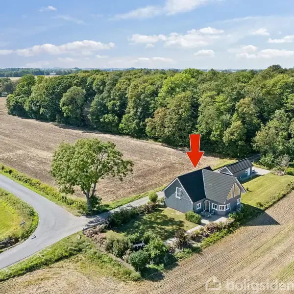 En hvid villa med sort tag står på en mark ved en vej og skov. Der er en rød pil over huset. Sidenavn er ikke relevant.