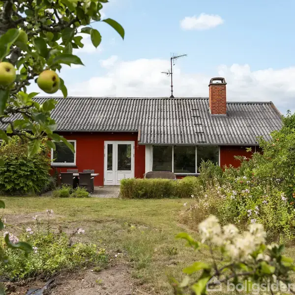 Rødt hus med gråt tegltag står i en have med frodige buske og et frugttræ med æbler. En terrasse med borde og stole ses foran huset under en blå himmel.