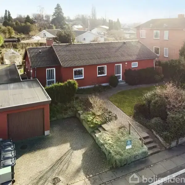 Rødt hus med fladt tag står ved siden af en garage, omgivet af en have med buske og træer. Flisebelagt sti fører til indgang. Skraldespande står til venstre langs indkørslen.