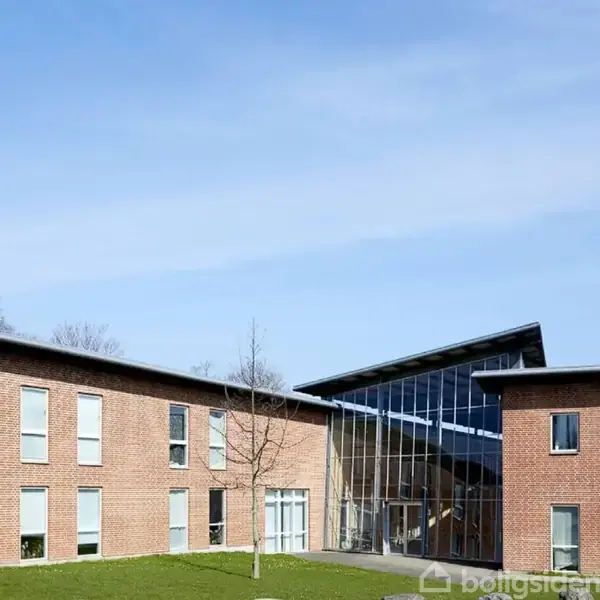 En moderne bygning med røde mursten og store glaspartier står stille under en blå himmel, omgivet af en grøn græsplæne.