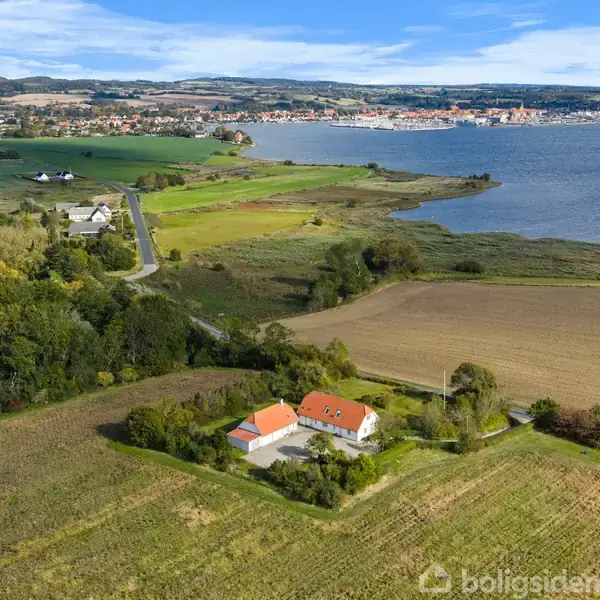 Hus med rødt tag står stille omgivet af frodige marker og tæt på en vej; i baggrunden ses en rum by ved siden af en stor sø under en blå himmel.