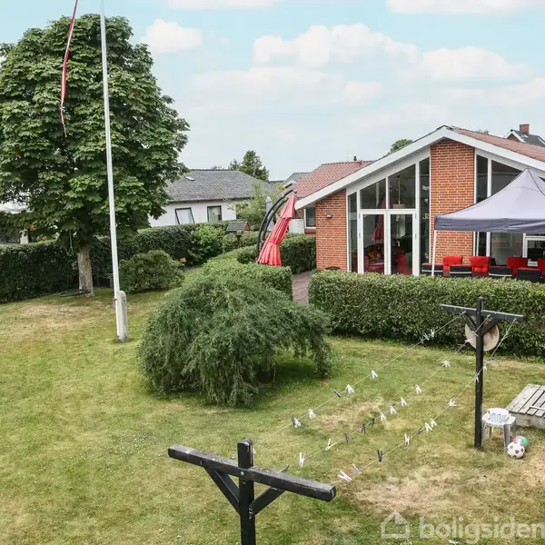 Rødt murstenshus med store vinduer, placeret i en velplejet have med græsplæne, træer, tørresnor med tøj, flagstang og et havebord med parasol.
