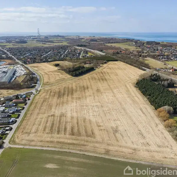 En stor, pløjet mark strækker sig i forgrunden mod havet i det fjerne. Omkring marken ligger huse og træer langs veje.